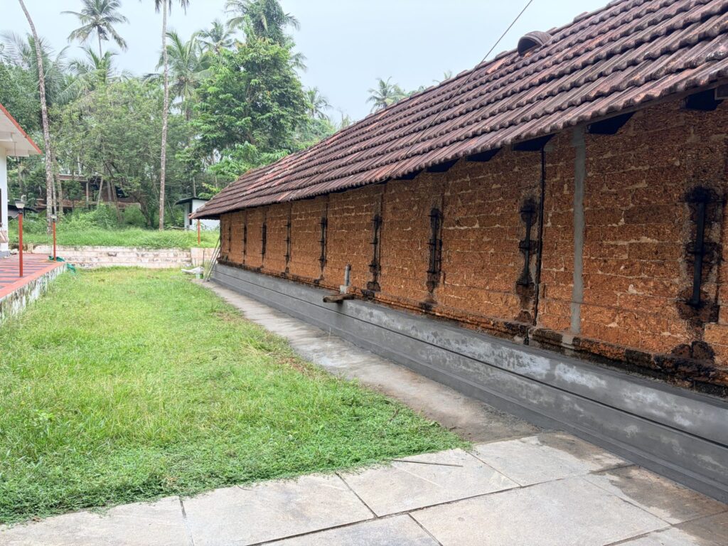 A long, traditional Kerala-style temple building features a weathered, sloped roof covered in reddish-brown clay tiles. The exterior wall is constructed from exposed, rough-textured red laterite stone blocks, punctuated by narrow vertical wooden windows with black iron grills spaced evenly along the side.