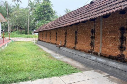 A long, traditional Kerala-style temple building features a weathered, sloped roof covered in reddish-brown clay tiles. The exterior wall is constructed from exposed, rough-textured red laterite stone blocks, punctuated by narrow vertical wooden windows with black iron grills spaced evenly along the side.