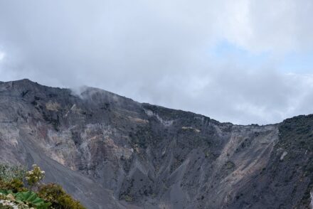 A panoramic view of a volcanic crater, featuring steep, rocky walls that rise sharply against a cloudy sky.