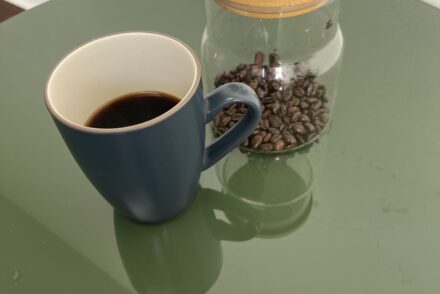 A dark blue cup of black coffee beside a glass jar of coffee beans on a green round table.