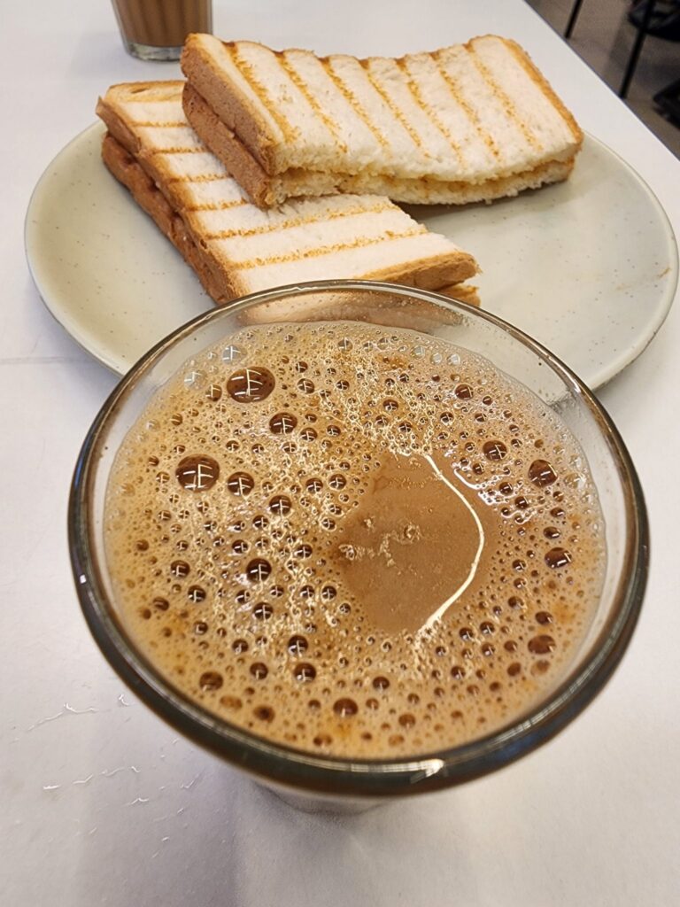 A glass filled with frothy, dark brown liquid, likely a coffee or chocolate drink, is placed prominently in the foreground. In the background, there’s a light-colored round plate holding several slices of grilled bread, which have a golden-brown crust and are arranged neatly. The slices appear to have been toasted with distinct grill marks, and some have been partially cut, revealing a soft, white interior. A blurred cup of similar drink is visible behind the plate, adding to the café setting. The surface beneath the items is white, creating a clean, simple backdrop.