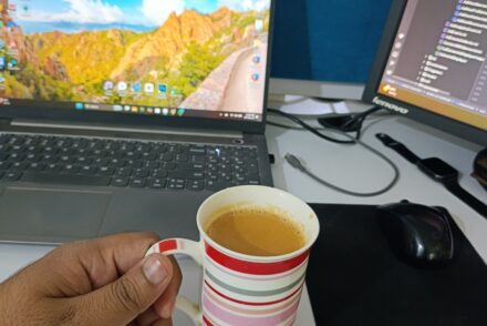 A hand holds a striped mug of tea at a white desk with a laptop and monitor, creating a focused workspace.