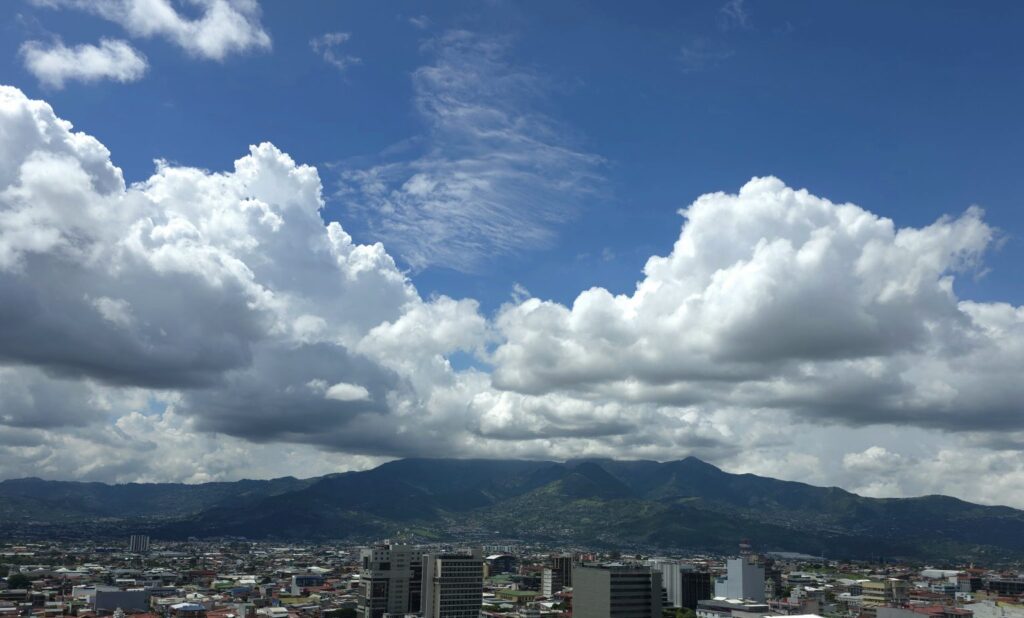 San José Skyline Beneath Mountain Clouds, Panoramic view of San José, Costa Rica, with the city skyline, surrounding mountains, and dramatic cloud formations rising above the Central Valley