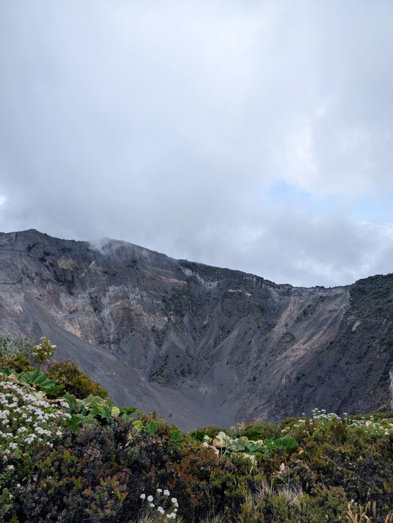 A panoramic view of a volcanic crater, featuring steep, rocky walls that rise sharply against a cloudy sky.