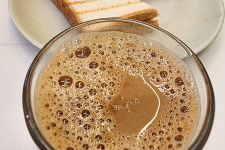 A glass filled with frothy, dark brown liquid, likely a coffee or chocolate drink, is placed prominently in the foreground. In the background, there’s a light-colored round plate holding several slices of grilled bread, which have a golden-brown crust and are arranged neatly. The slices appear to have been toasted with distinct grill marks, and some have been partially cut, revealing a soft, white interior. A blurred cup of similar drink is visible behind the plate, adding to the café setting. The surface beneath the items is white, creating a clean, simple backdrop.