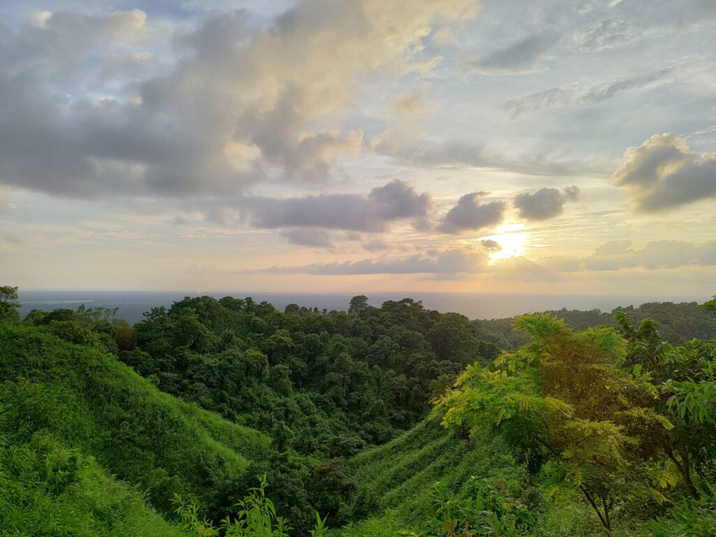 The setting sun glows through dramatic clouds above lush green forested hills, with the sea visible as a faint line on the far horizon.