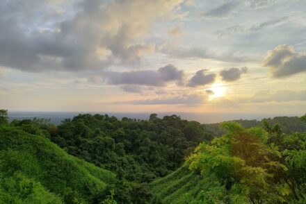 The setting sun glows through dramatic clouds above lush green forested hills, with the sea visible as a faint line on the far horizon.