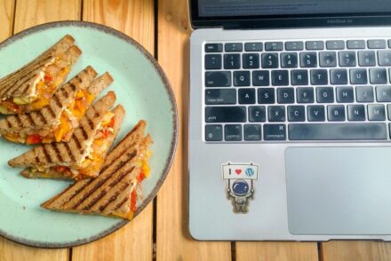 Grilled vegetable sandwiches served on a ceramic plate beside a laptop on a wooden table.