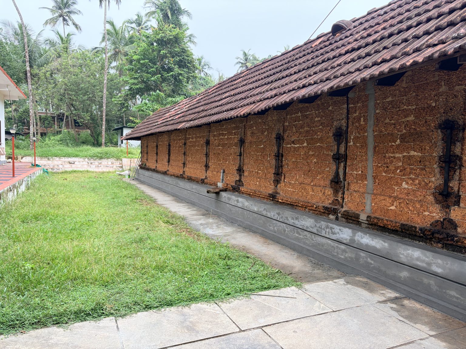 A long, traditional Kerala-style temple building features a weathered, sloped roof covered in reddish-brown clay tiles. The exterior wall is constructed from exposed, rough-textured red laterite stone blocks, punctuated by narrow vertical wooden windows with black iron grills spaced evenly along the side.