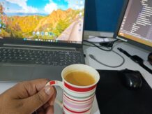 A hand holds a striped mug of tea at a white desk with a laptop and monitor, creating a focused workspace.