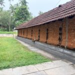 A long, traditional Kerala-style temple building features a weathered, sloped roof covered in reddish-brown clay tiles. The exterior wall is constructed from exposed, rough-textured red laterite stone blocks, punctuated by narrow vertical wooden windows with black iron grills spaced evenly along the side.
