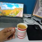 A hand holds a striped mug of tea at a white desk with a laptop and monitor, creating a focused workspace.